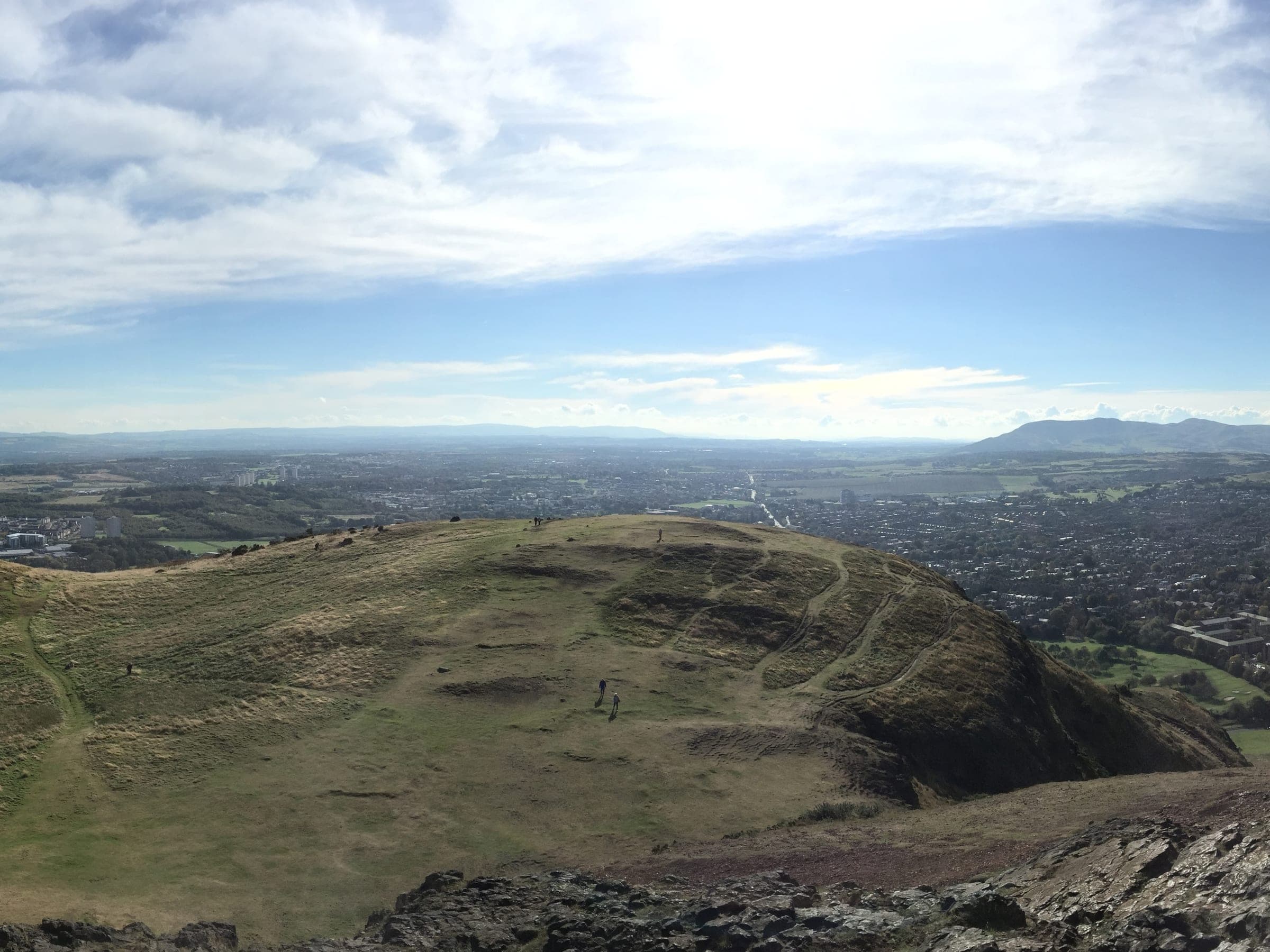 A grassy hilltop with a few walkers on it, looking out over a city to hills on the horizon.