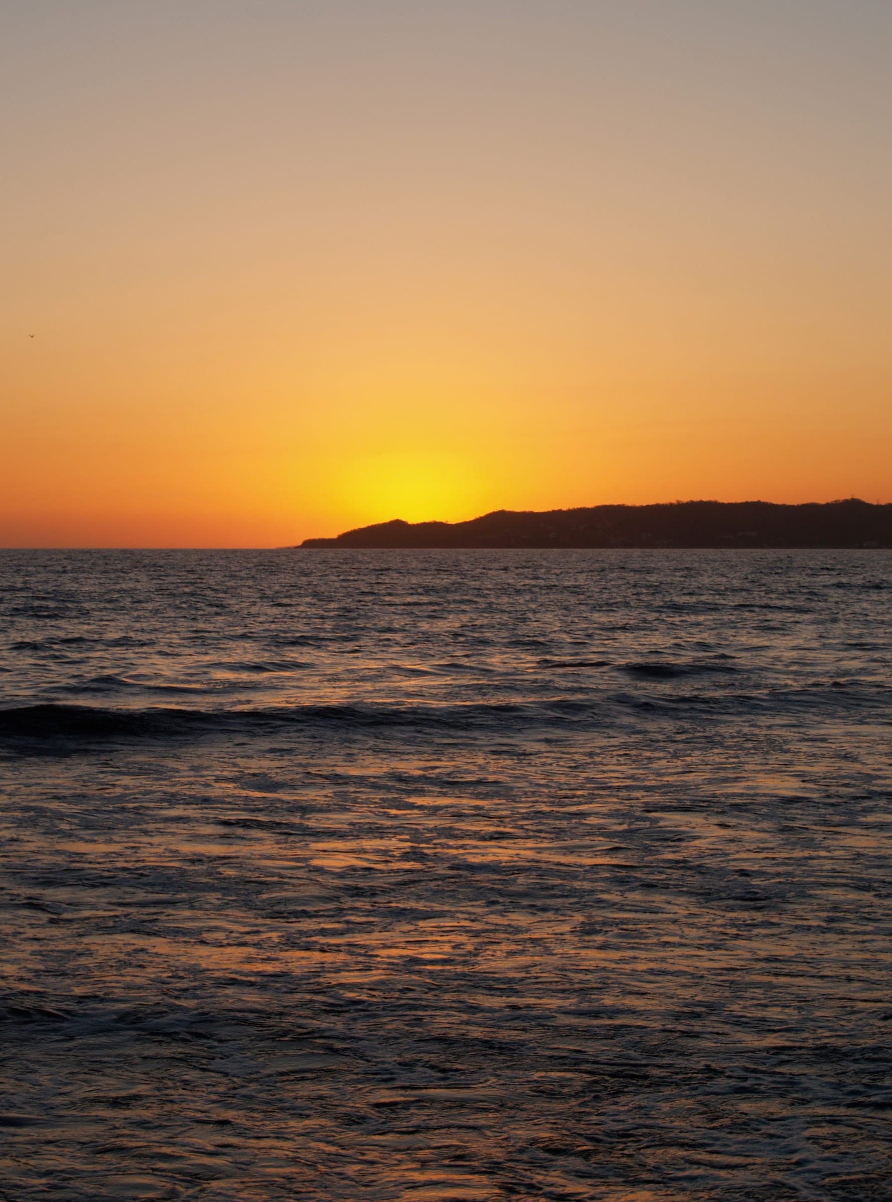 An orange sunset behind a low headland, over a calm sea.