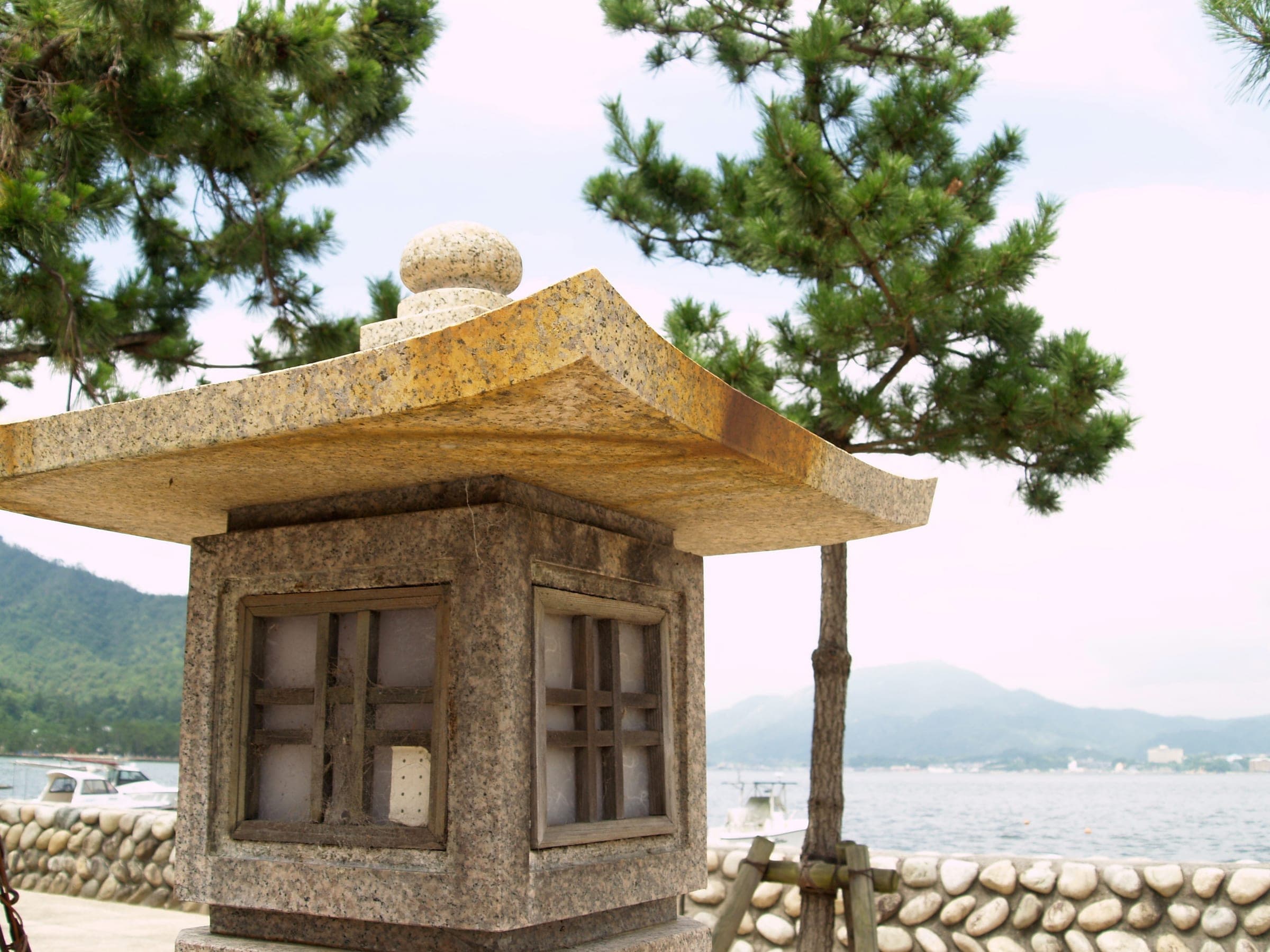 A stone lantern under pine trees by the shore, with hills across the water.
