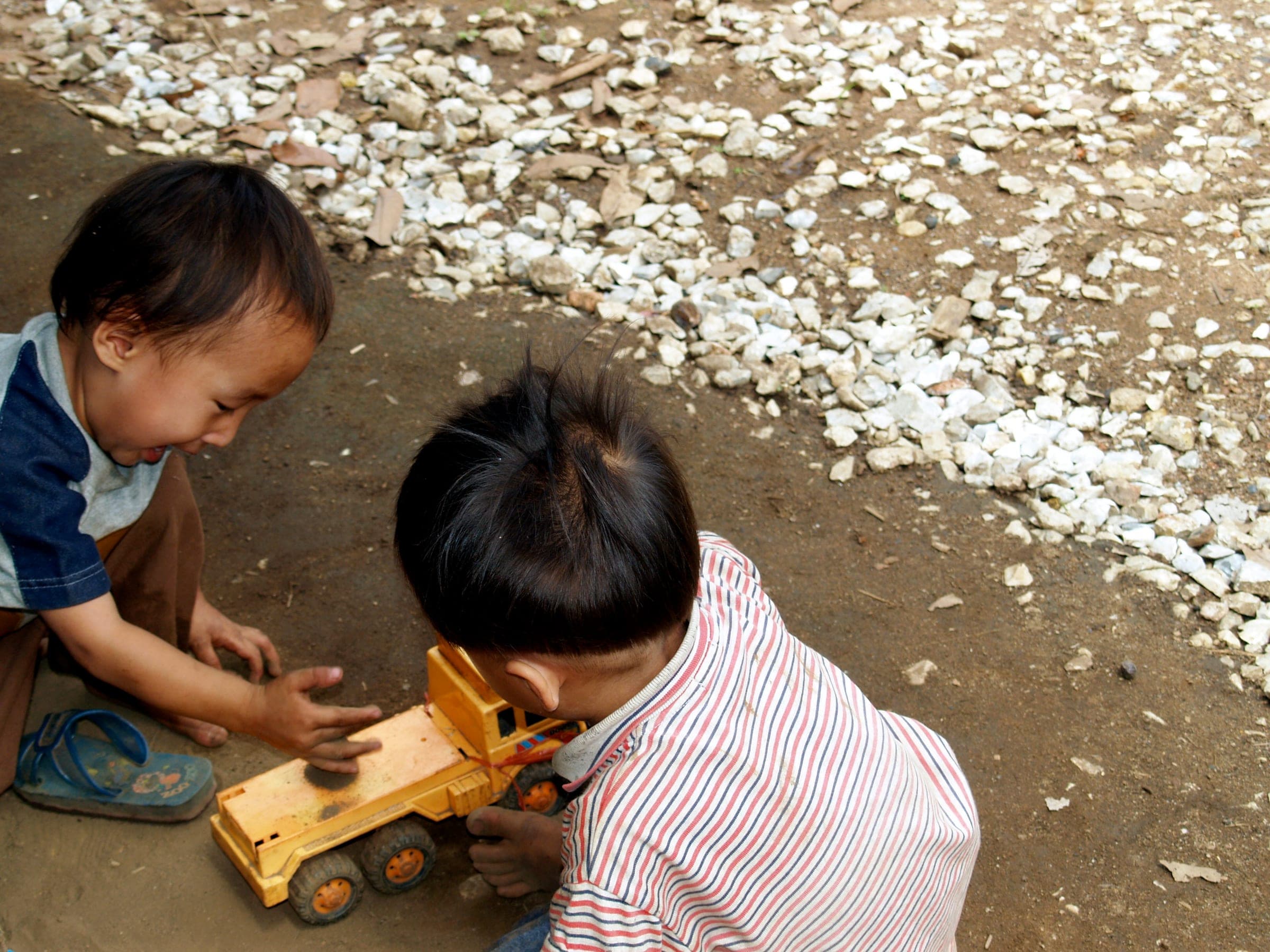 Two small children crouched over a yellow toy truck in the sand.